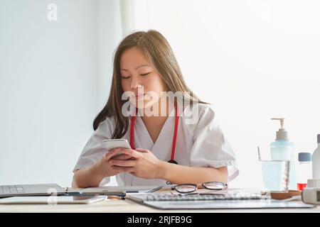 Concentrated Asian female therapist in uniform checking information on cellphone while sitting at table with documents during work in clinic Stock Photo
