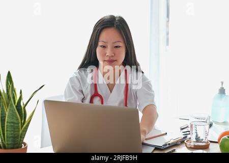 Concentrated Asian female therapist in medical uniform browsing modern netbook while sitting at table with documents during work in contemporary hospi Stock Photo