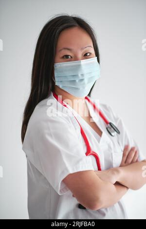 Side view of confident woman in medical uniform with stethoscope crossing arms and looking at camera on white background Stock Photo