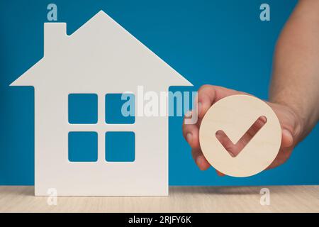 Real estate mortgage approval. Check mark icon in hand and house model as a symbol of a successful real estate purchase or sale deal. Photo with blue Stock Photo