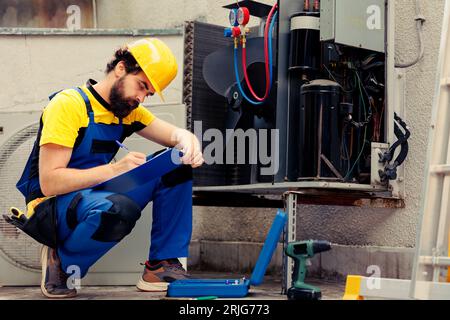 Qualified worker commissioned for outside air conditioner annual maintenance, looking for broken internal parts. Competent wireman doing hvac system inspection, writing report on clipboard Stock Photo