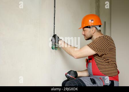 Electrician male in uniform, protective gloves and helmet checks presence of electrical voltage in socket phase uses electrical tester screwdriver.Exa Stock Photo