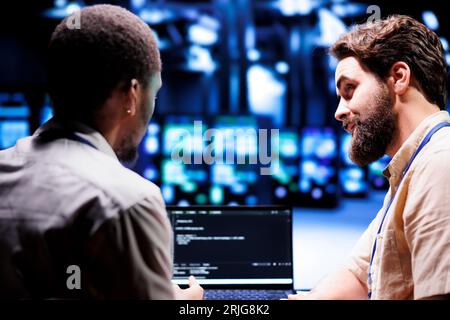 Coworkers updating blade servers, writing complex binary code scripts on laptop terminal. Employees using JavaScript, Java, and database programming to safeguard data using encryption techniques Stock Photo