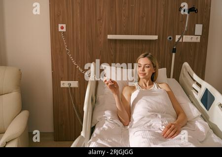 Young smiling woman patient holding emergency call button while lying in hospital bed Stock Photo