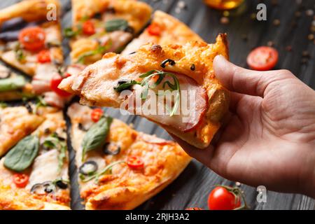 Hands take slices of cut pizza on a dark wooden background top view. Pizza with salami, jerky, sauces, spices, fresh cherry tomatoes on a black Stock Photo