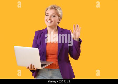 Happy young female programmer with laptop waving hand on yellow background Stock Photo