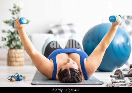 Overweight lady doing sports exercise in living room. He lies on the mat and lifts dumbbells. Stock Photo