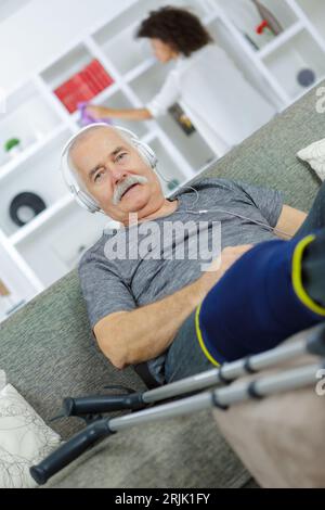 injured man listening to headphones woman doing chores in background Stock Photo
