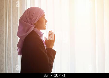 Muslim woman in pink hijab praying for life in apartment Stock Photo