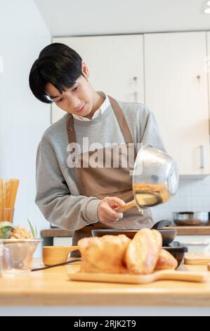 A happy and handsome young Asian man in an apron focuses on cooking, pouring batter on a pan, making pancakes in his minimalist kitchen. Stock Photo