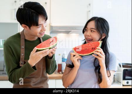 A lovely and joyful young Asian couple enjoys eating watermelon together in the kitchen, spending fun time together at home. Cooking date, romantic do Stock Photo