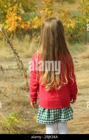 Beautiful little girl long blond hair in pink jacket plaid skirt on autumn nature background back view. Cute child walking alone in forest. Psychology Stock Photo