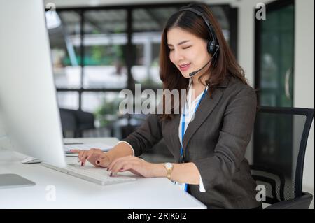 A professional and beautiful Asian female customer support agent is working on her computer, answering incoming calls and fixing customers problems on Stock Photo