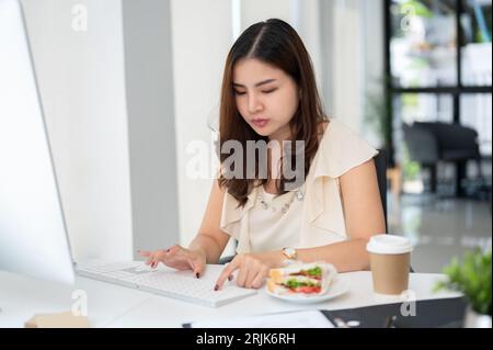 A hardworking Asian business woman is working on her assignment on computer while eating a sandwich at her table in the office. Stock Photo