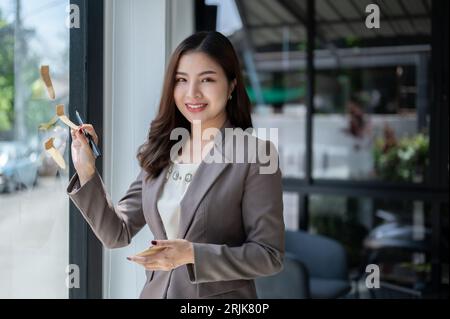 A beautiful and smiling millennial Asian businesswoman attached sticky notes to a glass wall in her office. creative ideas, memo notes, brainstorming Stock Photo