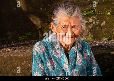 Ubud, Bali - July 29, 2016. Close-up of a very elderly Balinese woman smiling while sitting outdoors in a garden. Stock Photo