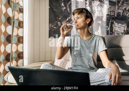 Young blonde drinks water from a glass sitting at home with a laptop. Stock Photo