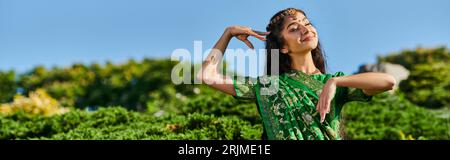 cheerful indian woman in matha patti and sari posing near plants with blue sky on background, banner Stock Photo
