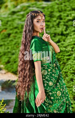 young indian woman in sari showing secret gesture and looking at camera near plants in park Stock Photo