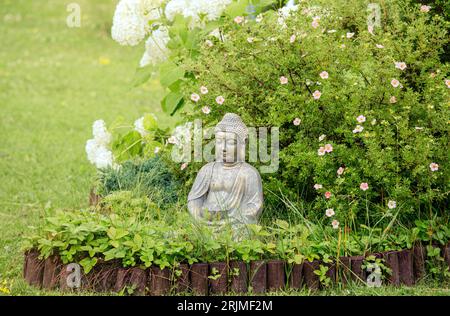 Meditating Buddha figure sit inside small island of flowers in flower bed, surrounded by natural wood roll palisade in home garden in summer. Stock Photo