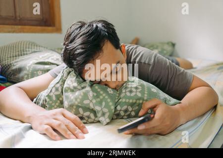 Portrait of Asian man fall asleep while holding smartphone laying down on bed Stock Photo