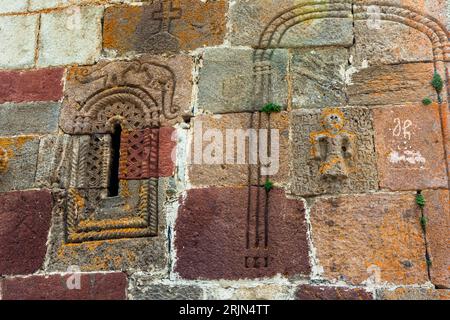 Stone reliefs decorate an exterior wall of Gergeti Trinity Church (Georgian გერგეტის სამების ეკლესია) near Stepantsminda (Georgian სტეფანწმინდა, literal translation of the Georgian name: Holy Stephen), Mtskheta-Mtianeti region, Georgia. A narrow window sits in a frame of interlace ornament beneath a round arch. Above the arch, a relief shows an animal with a long, curled tail. A cross is carved above it. On the right, a block bears a small human figure surrounded by carved Georgian characters. Two moulded arches run across the upper right. The blocks are grey, reddish and yellowish, with orang Stock Photo