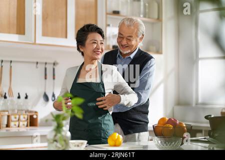 loving senior asian man husband helping wife tying up apron in kitchen at home Stock Photo