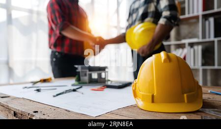 Yellow safety helmet on workplace desk with construction worker team hands shaking greeting start up plan new project contract in office at Stock Photo