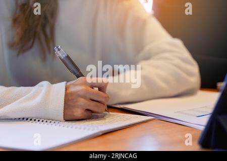 A closeup of a university student writing on notebook with tablet on the table Stock Photo