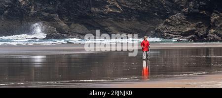 A panoramic image of mature female wearing a bright red coat and using walking poles hiking sticks walking on the shore at Mawgan Porth beach in Cornw Stock Photo