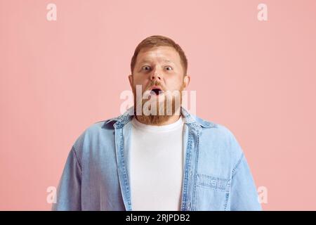 Bearded man in casual clothes standing with wide open eyes and mouth, showing shocked face against pink studio background Stock Photo
