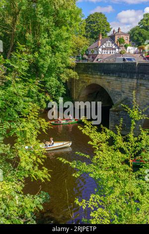 Tourists in two rowing boats paddle towards an arch bridge in Knaresborough, framed by lush trees and the World's End Pub, England, UK. Stock Photo