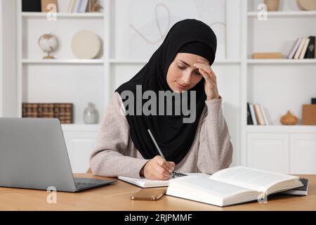 Tired Muslim woman in hijab working near laptop at wooden table in room Stock Photo
