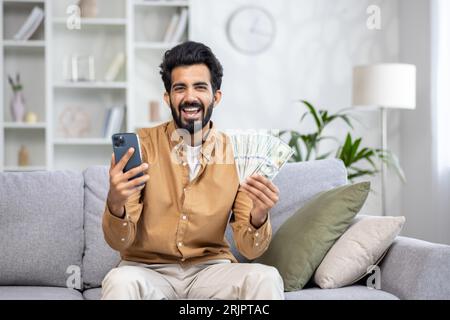 Male winner of online game sitting on sofa at home, hispanic joyfully shouting and smiling looking at camera, holding phone and money cash dollars, using app on smartphone. Stock Photo