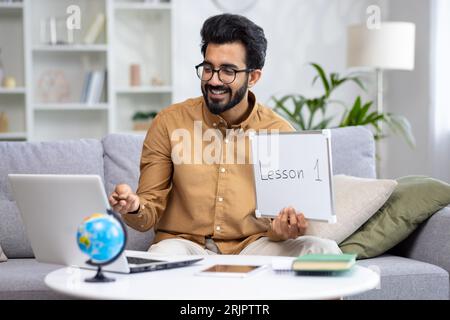 Young male teacher teaching remotely from home, hispanic using laptop for video call and communication and teaching students remotely, explaining new school material smiling. Stock Photo