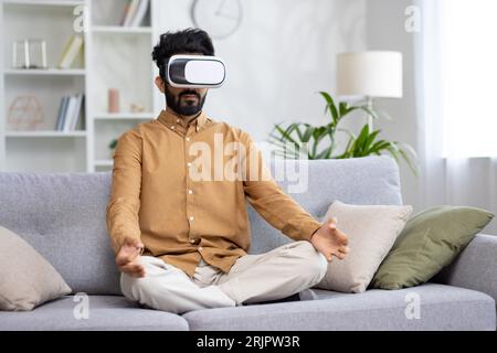Young hispanic man meditating at home sitting on sofa, using vr virtual reality glasses, sitting in lotus position. Stock Photo