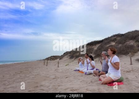 Group of men and women practicing kundalini yoga on the sand at the beach in front of the sea. Abhaya pose Stock Photo