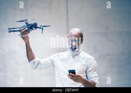 Portrait of bald mature man holding quadcopter while looking at cell phone Stock Photo