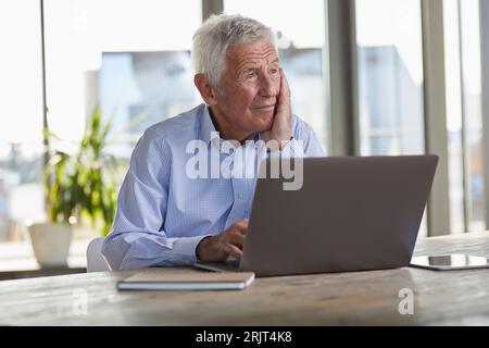 Portrait of pensive senior man sitting at table with laptop looking at distance Stock Photo