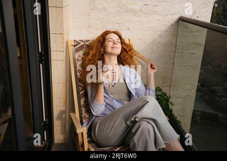 Smiling woman sitting on chair holding glass of water in balcony Stock Photo