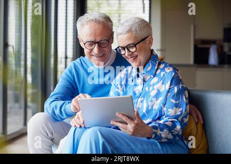 Happy senior couple sitting on couch at home using tablet PC Stock Photo