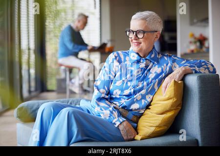 Relaxed senior woman sitting on couch at home with man in background Stock Photo