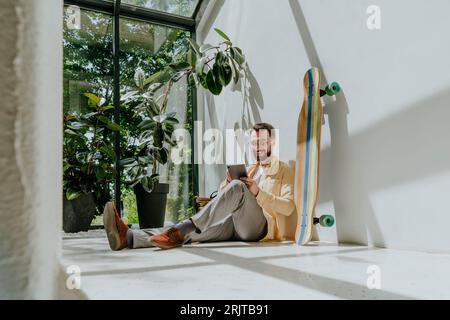 Smiling businessman sitting by longboard and using tablet PC in office Stock Photo