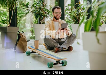 Happy businessman sitting with longboard and using tablet PC amidst plants in office Stock Photo