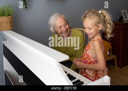 Smiling grandmother playing piano with granddaughter at home Stock Photo
