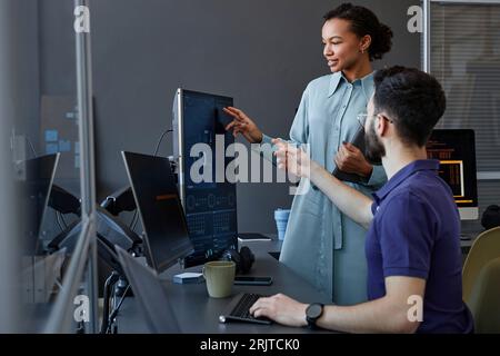 Smiling computer programmer gesturing and explaining to colleague over desktop PC Stock Photo