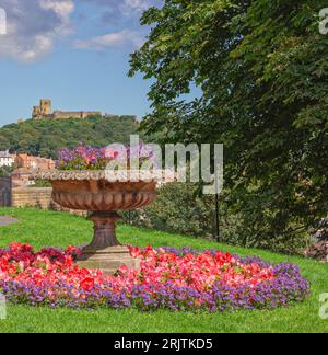 An ornamental flower pot is surrounded by a circle of red and purple flowers in a lawn. Houses are in the distance and a castle ruin stands atop a tre Stock Photo