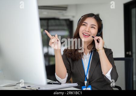 A cheerful and professional Asian female customer support agent or call centre operator is providing a positive customer exaperience over the call, gi Stock Photo