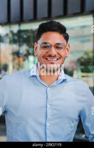 Vertical portrait of hispanic young entrepreneur smiling and looking at camera at workspace. Front view of one latin happy employee office worker with glasses and successful attitude standing outdoors. High quality photo Stock Photo