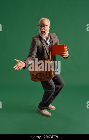 Full length portrait of senior man with beard in glasses, teacher wearing vintage suit with stack of books and brown business bag looks surprised Stock Photo